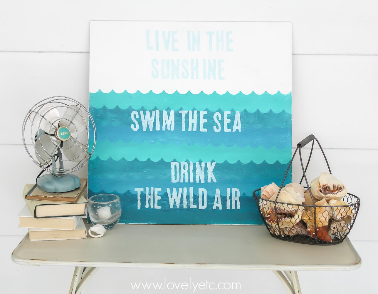 The perfect painted sign to welcome summer - so much easier to DIY than you would think! Live in the sunshine, swim in the sea, drink the wild air - Ralph Waldo Emerson. wooden sign with handpainted waves next to basket of shells.