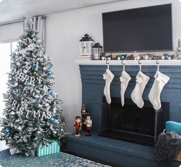 Christmas tree with blue and white ornaments next to dark blue fireplace with white stockings.