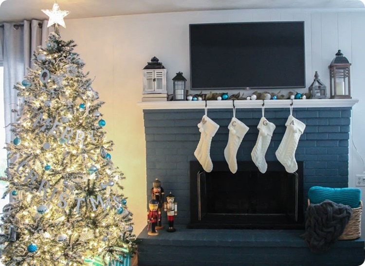 Flocked Christmas tree with blue and white ornaments next to fireplace with white stockings.