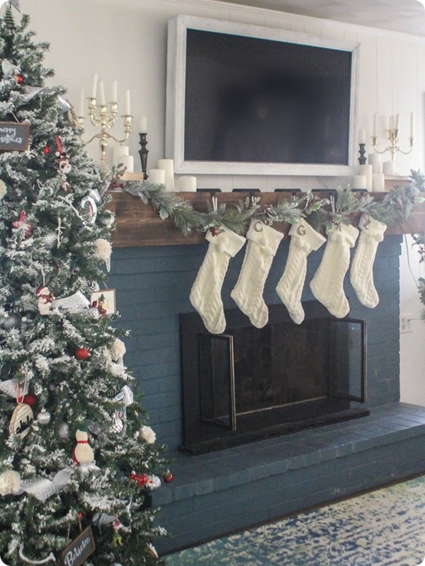 Blue brick fireplace with chunky wood mantel, flocked garland, white stockings, and candles around the TV.