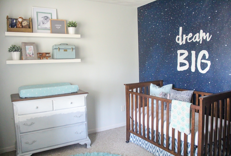 nursery with starry wall mural that reads Dream Big, crib, and dresser.