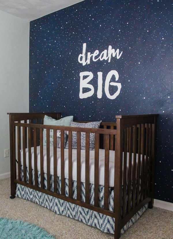 nursery with mural behind crib of a dark navy sky with white stars and big white letters that say Dream Big.