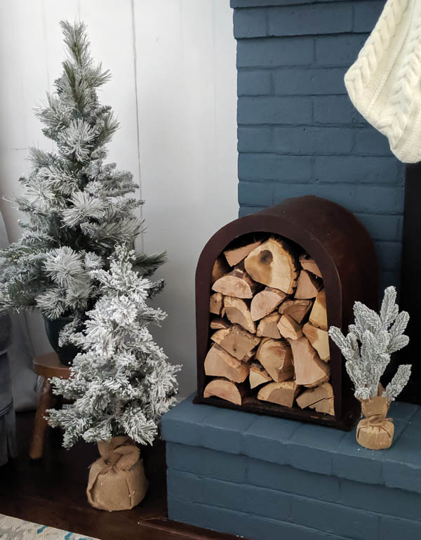 Cozy neutral Christmas fireplace with flocked Christmas trees, a unique firewood holder, and pretty white knit stockings.