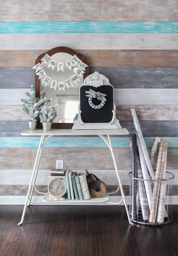 Entryway with weathered wood plank wall and white table with old mirrors, Merry Christmas sign, and mini Christmas trees next to basket of wrapping paper.