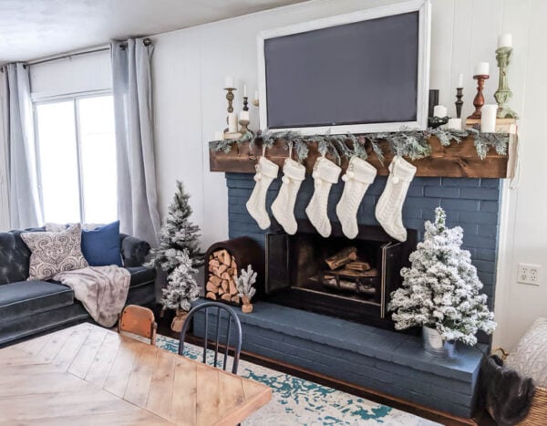 Brick fireplace painted blue with chunky wood mantel, garland, small flocked trees, and lots of candles.