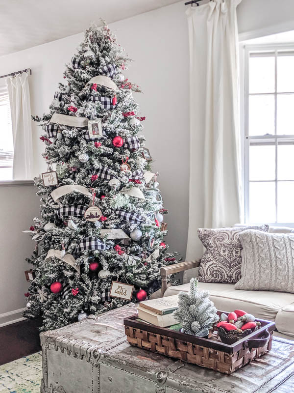 Classic Farmhouse Christmas tree with buffalo plaid ribbon, red berries and pinecones, and lots of handmade farmhouse ornaments next to a basket on the coffee table with a mini Christmas tree and bowl of glittered Christmas bulbs.