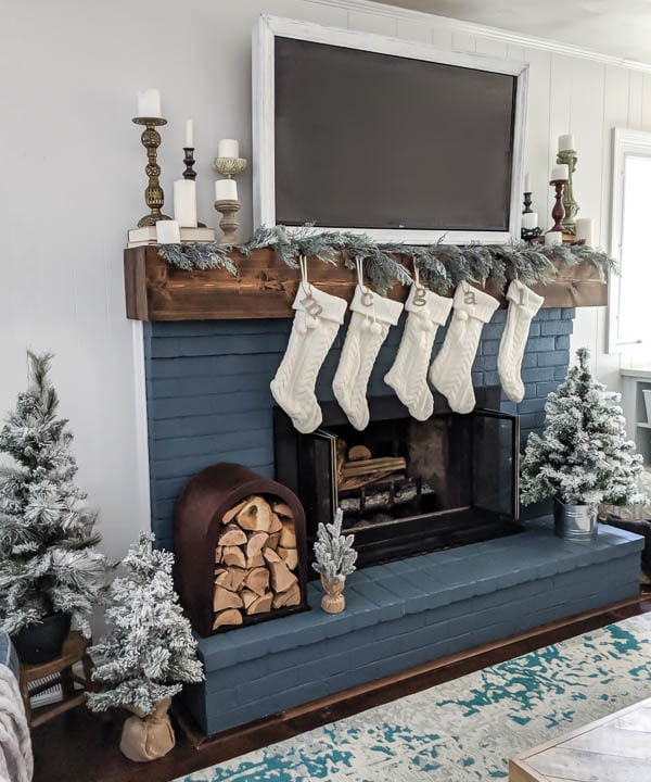 Garland, white Christmas stockings, pillar candles, and mini Christmas trees around brick fireplace.