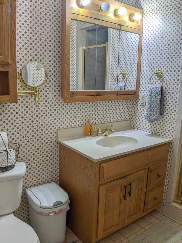 dated bathroom with busy wallpaper, oak vanity, huge oak medicine cabinet with attached light, and gold fixtures