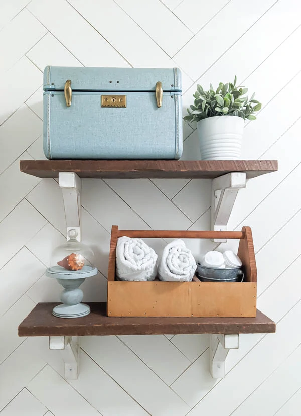 Open shelving over toilet with a small suitcase and wooden tool tote for storage and a small plant and cloche to accessorize.