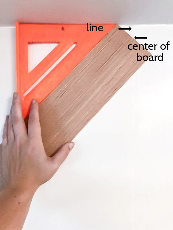 piece of wood held at a 45 degree angle to the ceiling next to a speed square with arrows pointing to the center of the board and the line marking the center of the wall