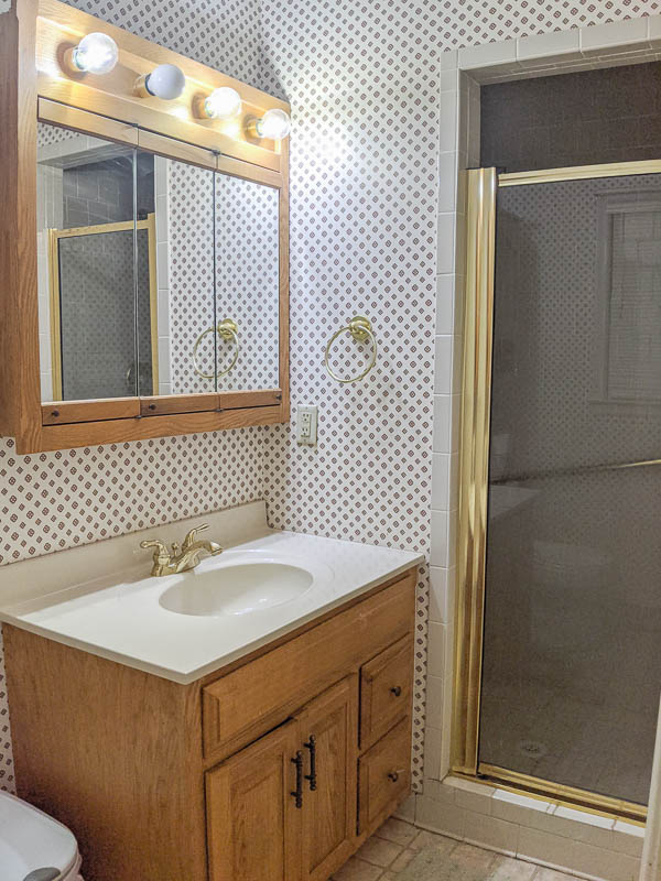 dated bathroom with wallpaper, a large oak vanity, a huge medicine cabinet and gold hardware