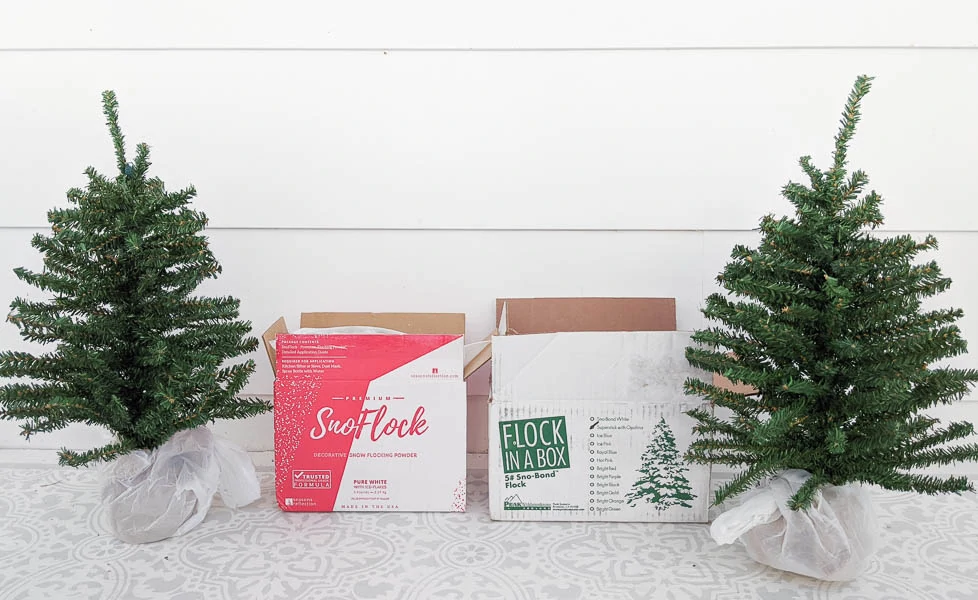 Two mini Christmas trees next to a box of SnoFlock and a box of Sno-Bond Flock in a Box snow flocking powder.