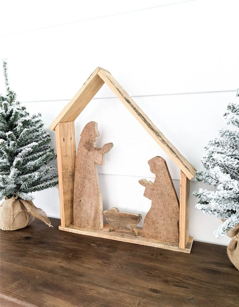 Simple DIY wood nativity set with stable, Jesus, Mary, and Joseph against a white shiplap wall flanked by two mini christmas trees.