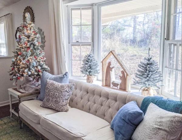 DIY wood nativity and small Christmas trees in bay window behind couch. 