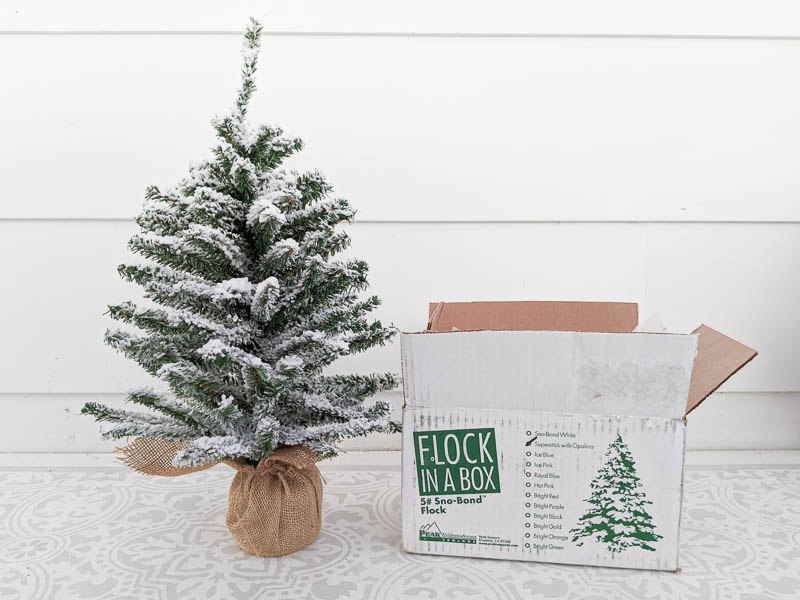 Small flocked Christmas tree next to a box of Flock in a Box snow flocking powder.