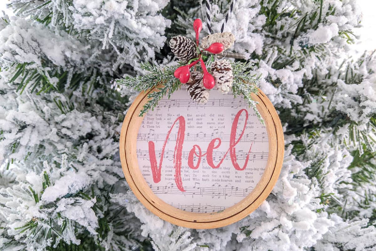 A round embroidery hoop ornament printed with the word Noel on sheet music, trimmed with evergreen on a snowy tree