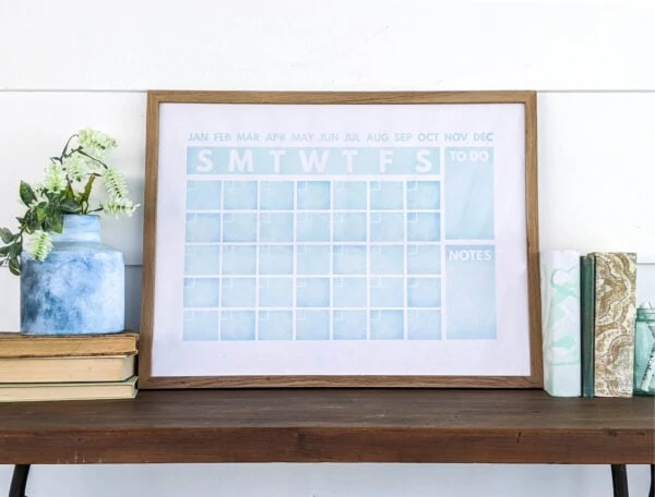light blue watercolor calendar framed in a wood frame and leaning against the wall on top of a desk.
