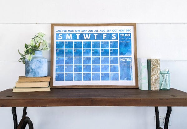 navy watercolor calendar framed in a wood frame and leaning against the wall on top of a desk.