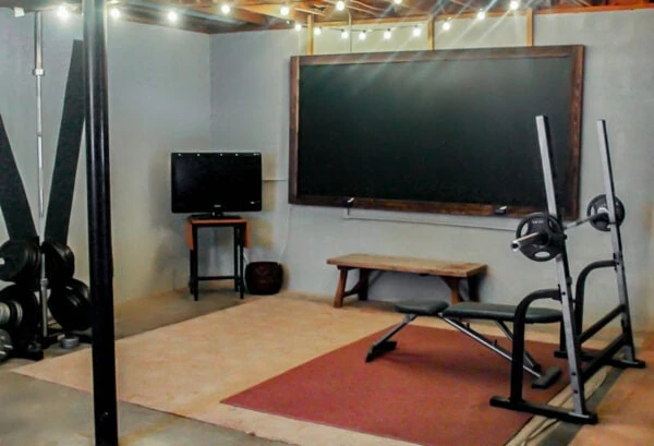 industrial basement gym with free weights, a TV, a rustic bench, and an oversized chalkboard.