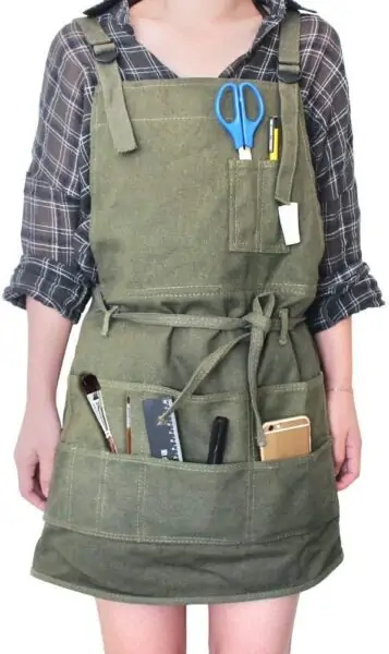 person wearing olive green canvas apron with scissors, paint brushes, and other supplies tucked in the pockets.