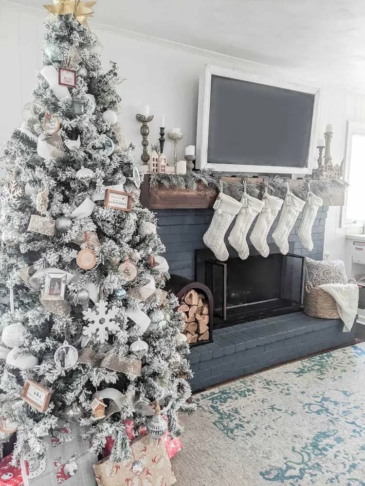 Christmas tree decorated with gold, white, and neutral ornaments next to blue brick fireplace with wood mantel and white stockings.