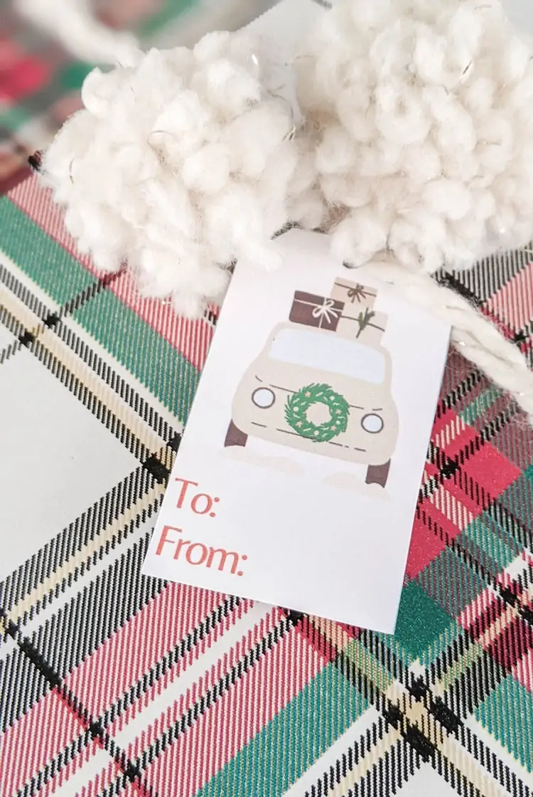 Christmas present wrapped in plaid paper with fuzzy pom poms and gift tag with cream car with wreath on front and presents on top.