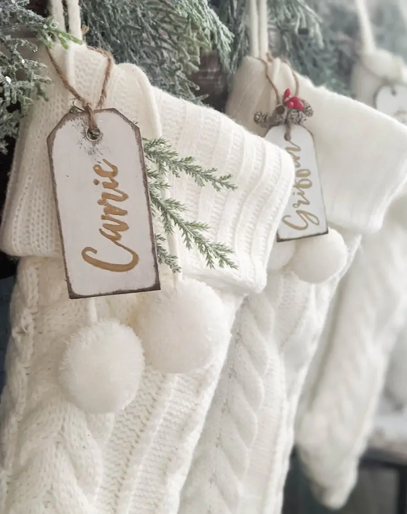 White stocking hanging on fireplace with evergreen garland and diy white and wood stocking name tags with gold script writing.