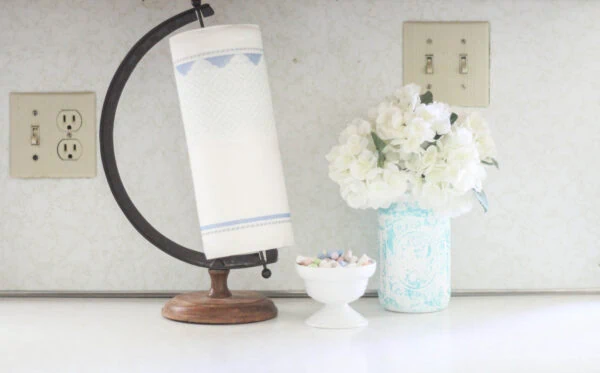 kitchen counter with paper towel holder and flowers in front of dingy beige electrical outlets and light switches.
