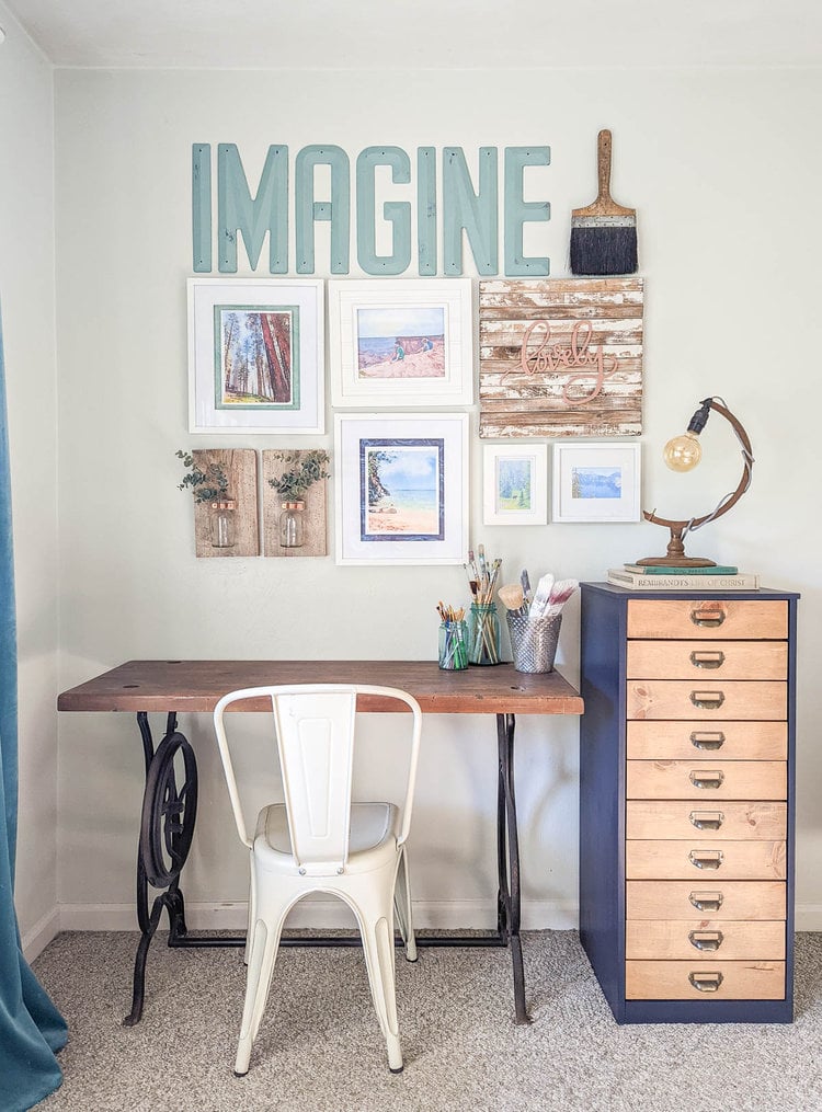 upcycled desk with gallery wall above with white framed photos, vintage letters spelling Imagine, a huge paintbrush, and some DIY wood pieces.