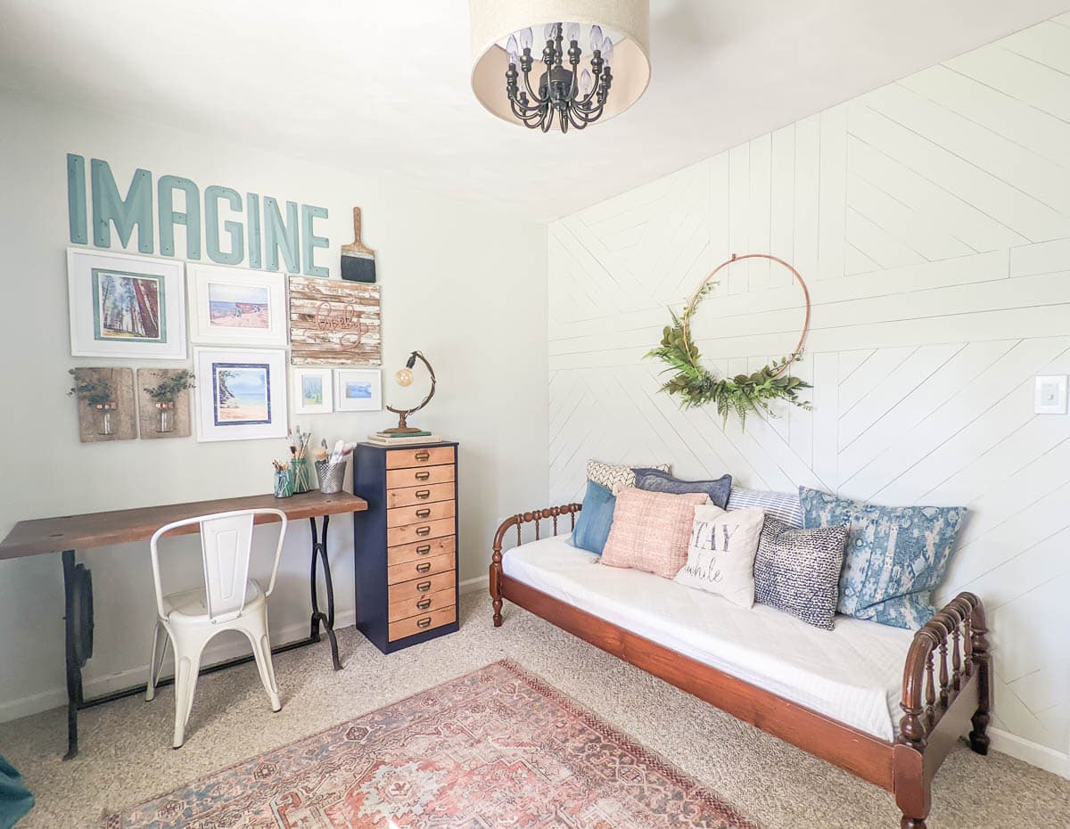view of two walls of office with wood wall and daybed on one wall and upcycled desk, chest of drawers and gallery wall on the adjoining wall.