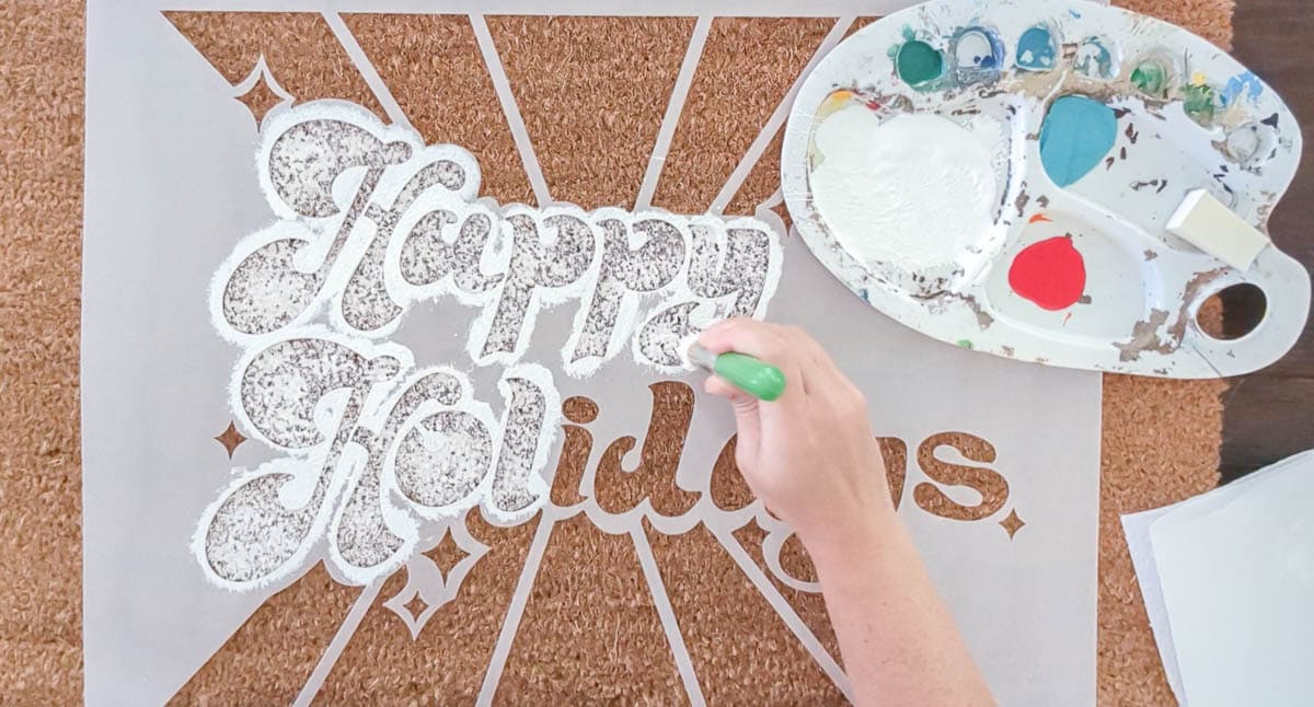using a stencil brush to paint the words happy holidays with white outdoor paint.