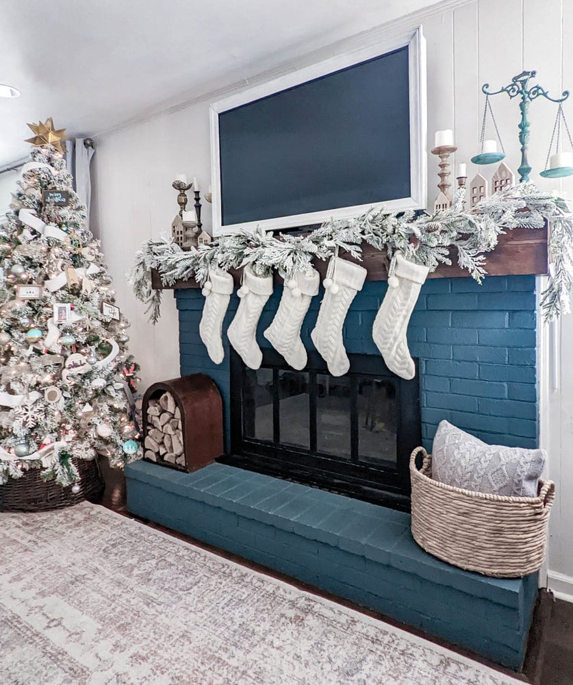 Dark blue brick fireplace decorated for Christmas with garland and white stockings next to flocked Christmas tree.