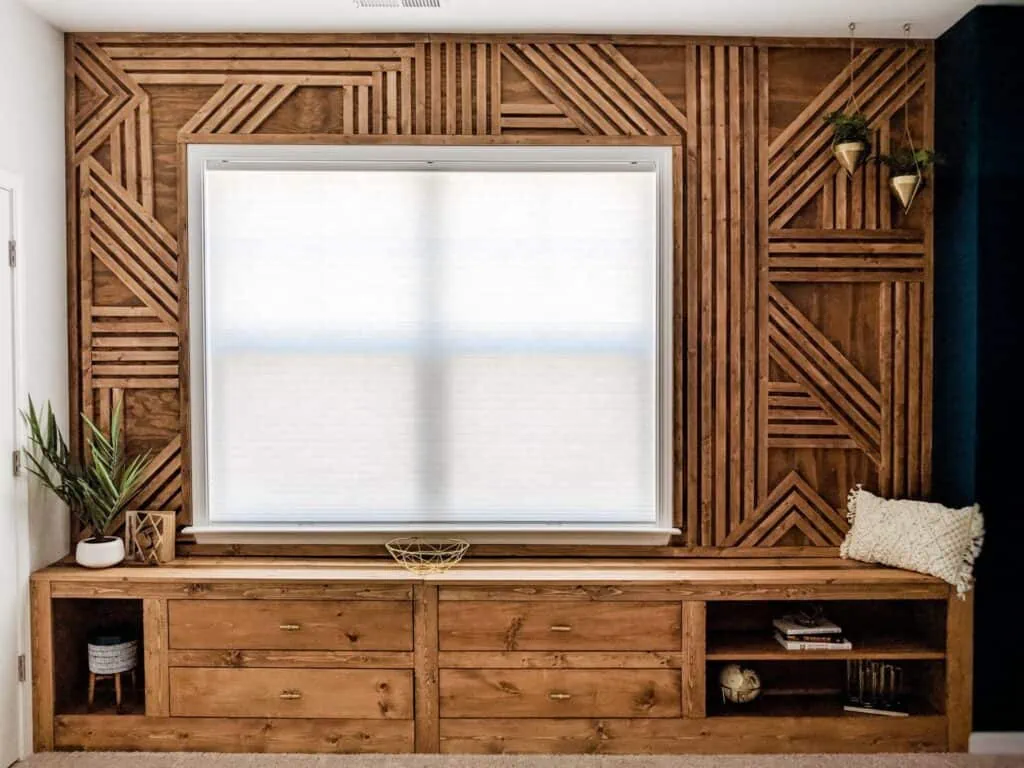 Wood accent wall made using plywood and 1 inch by 2 inch wood planks to create a 3D geometric wood wall around a window.