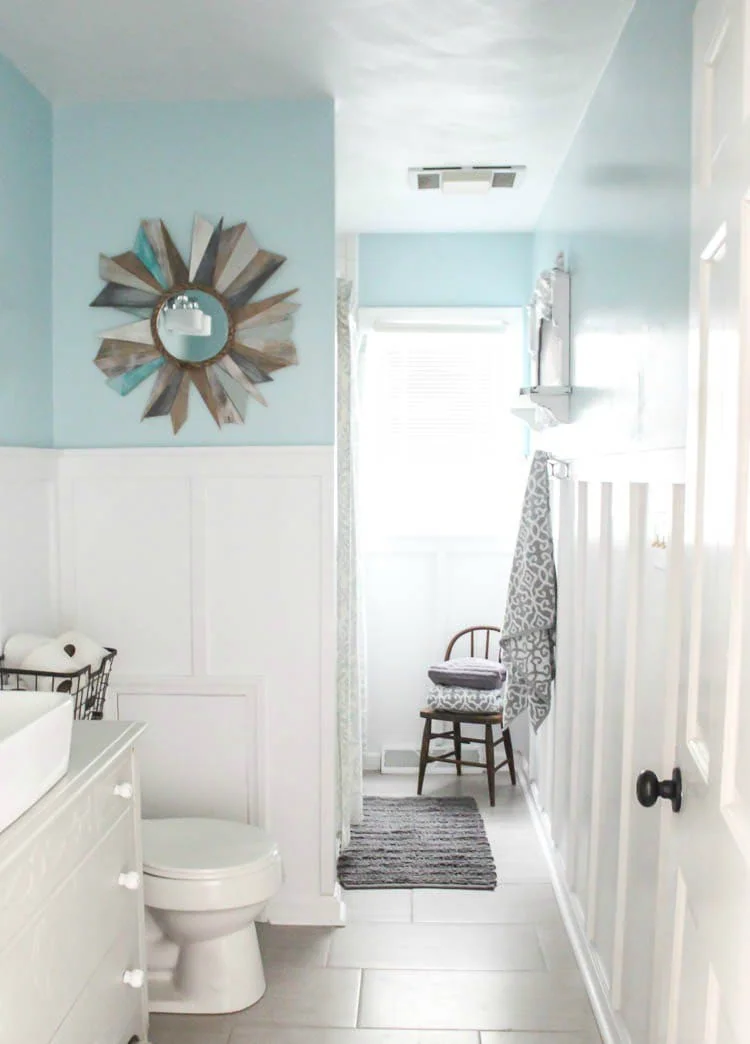 modern farmhouse bathroom with white board and batten walls with blue above and a wood sunburst mirror.