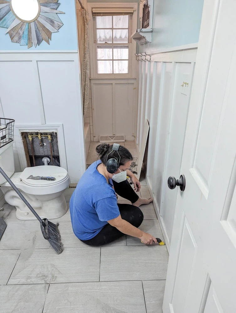 Carrie sitting in floor removing old grout while wearing face mask and ear protection.