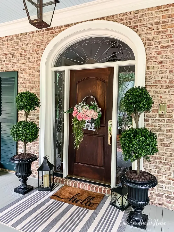 wood front door flanked by two large topiaries with a doormat that says hello layered over a striped rug.