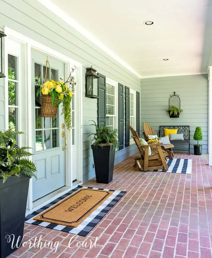 large front porch with tall planters with ferns on either side of front door and a welcome mat layered over a plaid rug on a brick floor.
