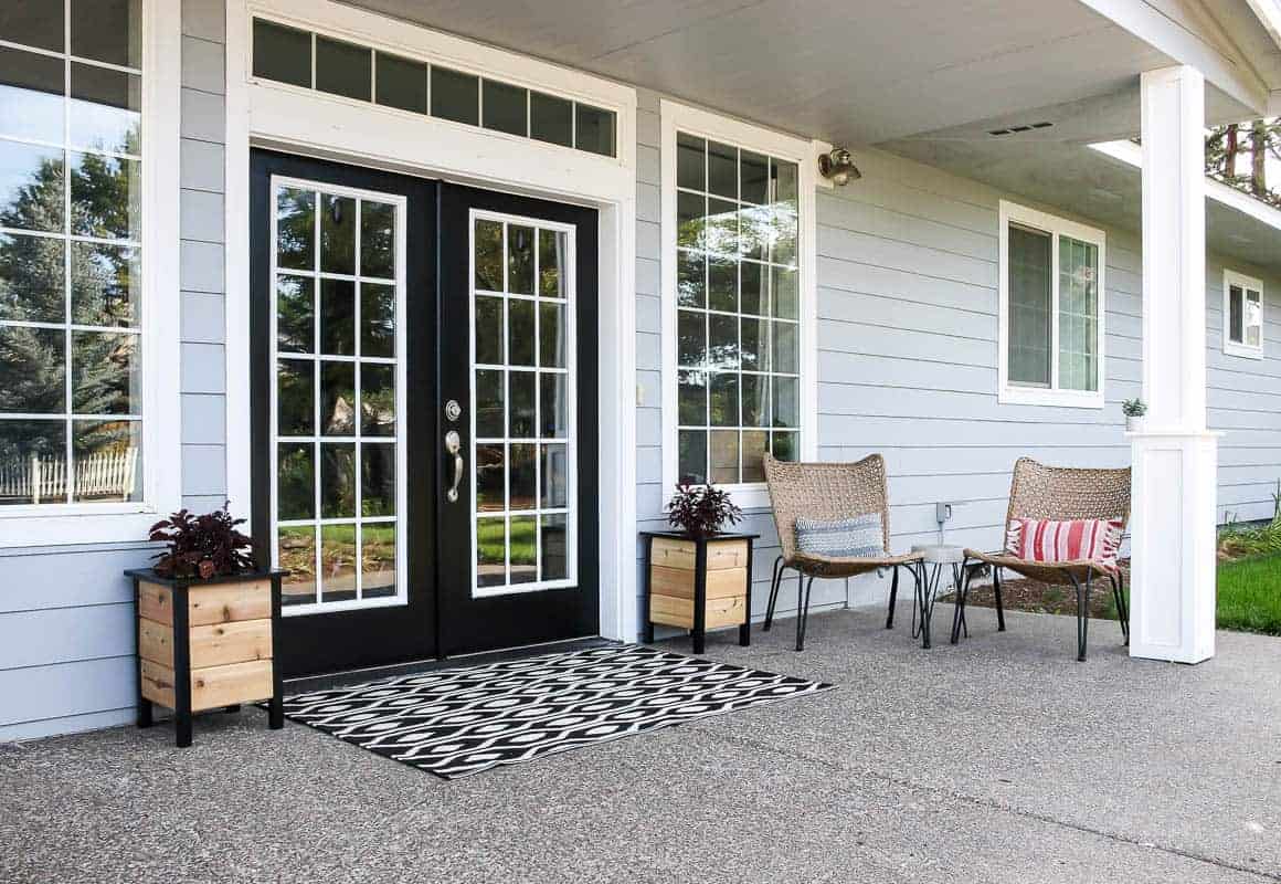 wide porch with black front door with wood plank planters on either side and a black and white rug in front.