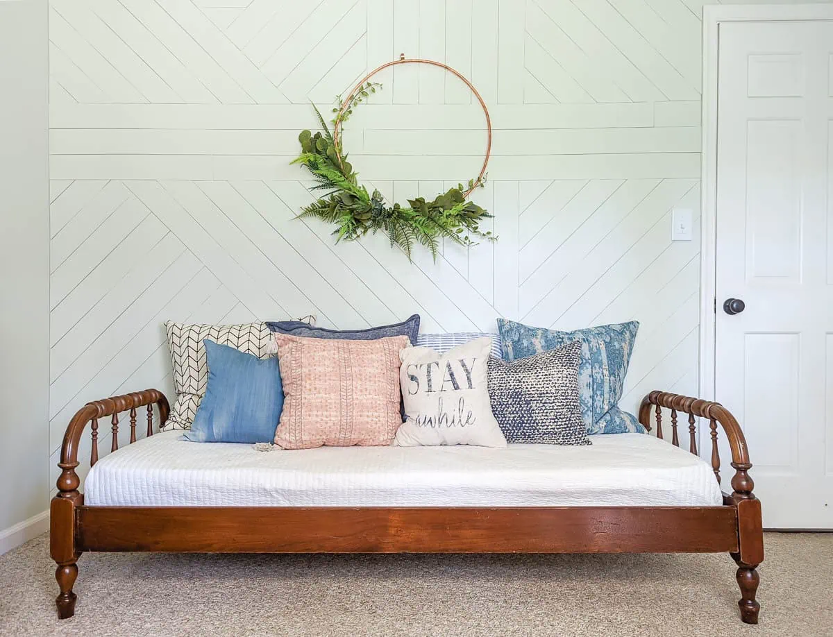 geometric wood accent wall painted white. With a large copper hoop wreath hanging on it and a wooden daybed in front.