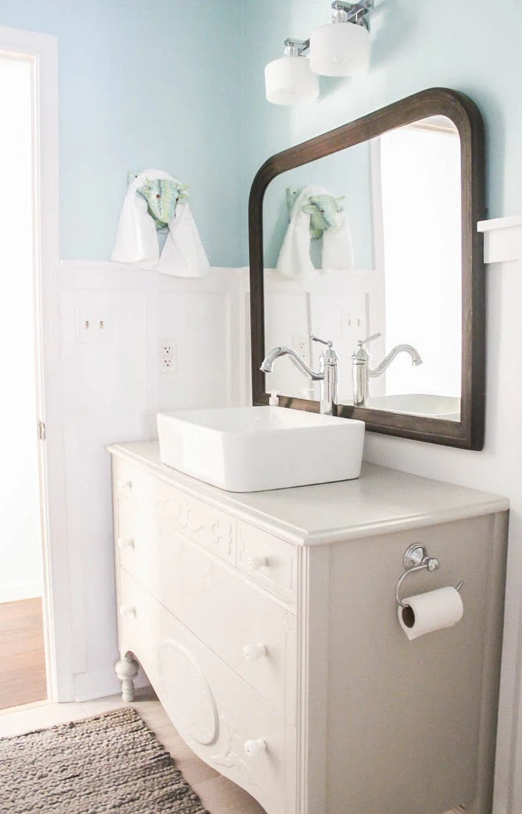 antique dresser bathroom vanity in bathroom with blue walls and white board and batten plus a vintage wooden arched mirror.