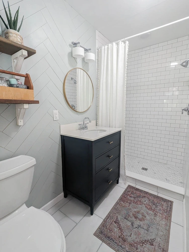 Master bathroom after makeover with subway tile walk in shower, navy vanity, gold round mirror, and marble look floor tile.