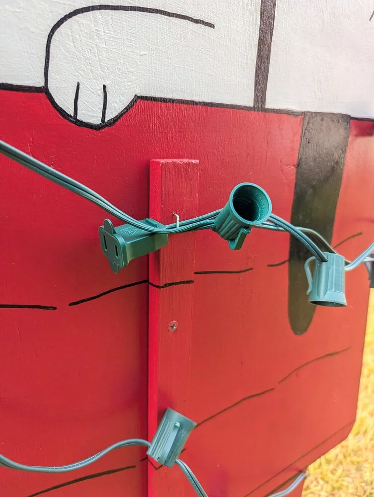 attaching Christmas lights to the outdoor wood cutout of Snoopy on his doghouse with a staple gun.